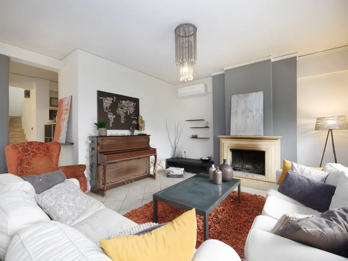 A living room with a white sofa, piano, fireplace, and orange rug. A chandelier hangs from the ceiling.