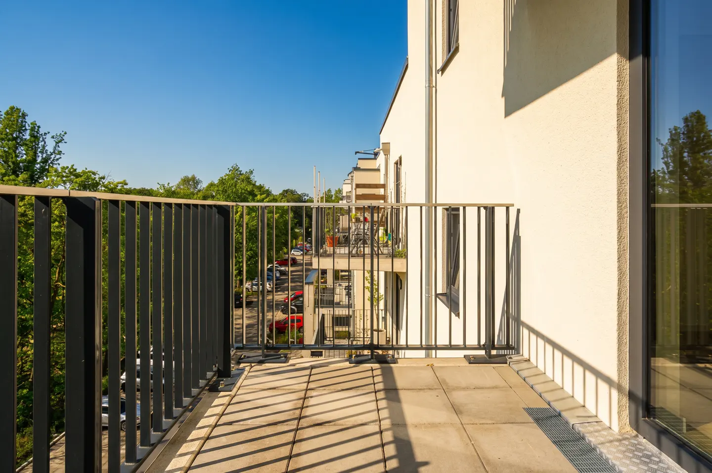 Apartment balcony with black metal railings and beige tile floor overlooking trees and a parking lot on a sunny day.