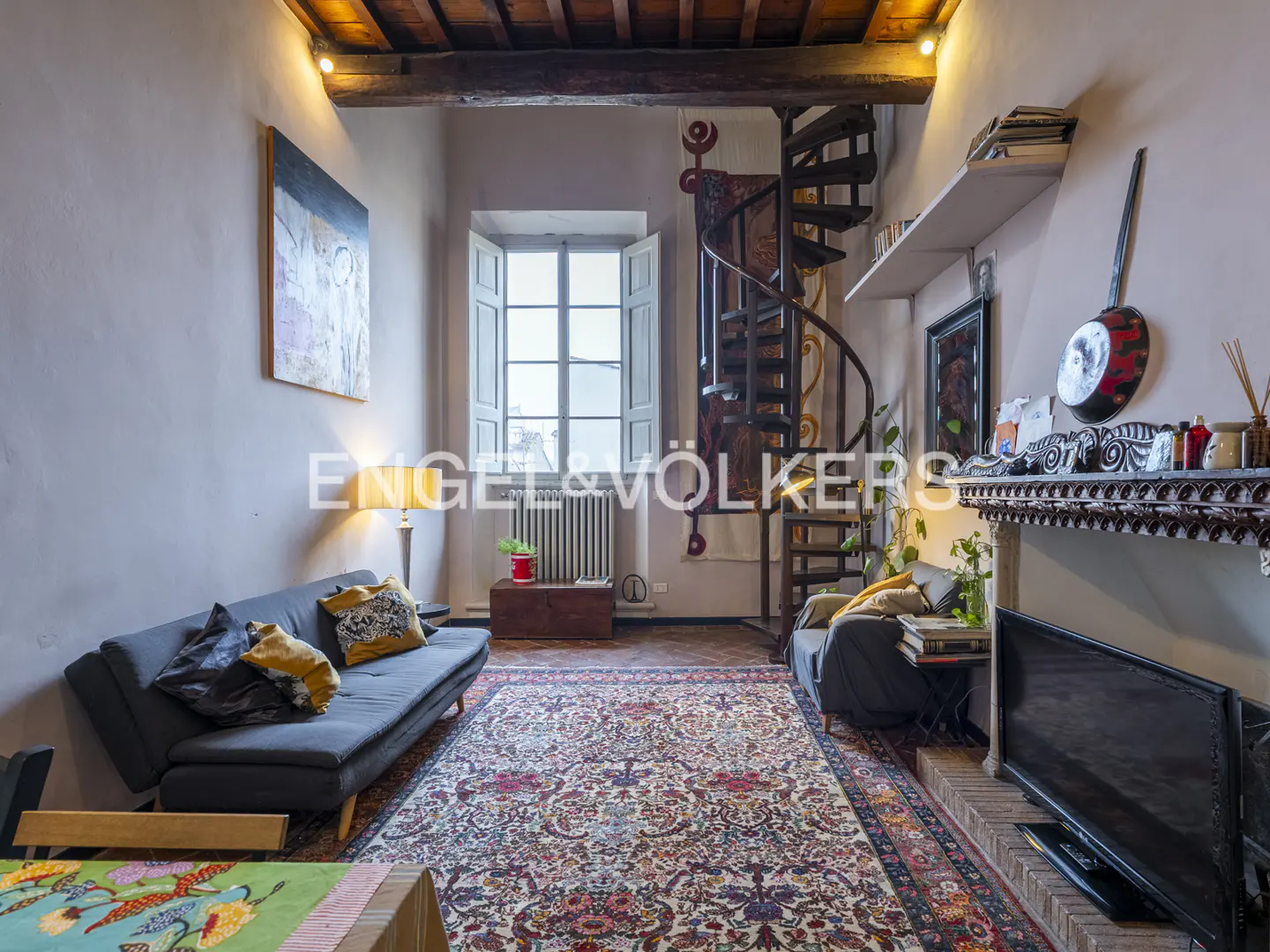 Living room with a blue sofa, a spiral staircase, and a patterned rug. A fireplace with a TV is on the right.