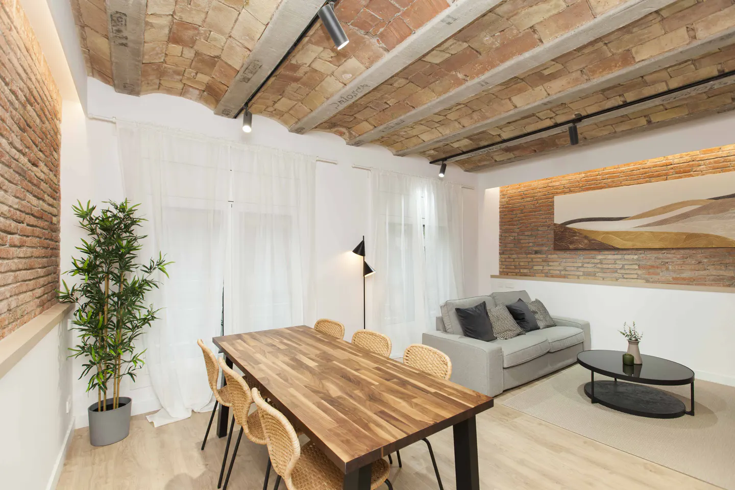 Bright living room with brick vaulted ceiling, track lighting, wood table with wicker chairs, and a gray sofa.