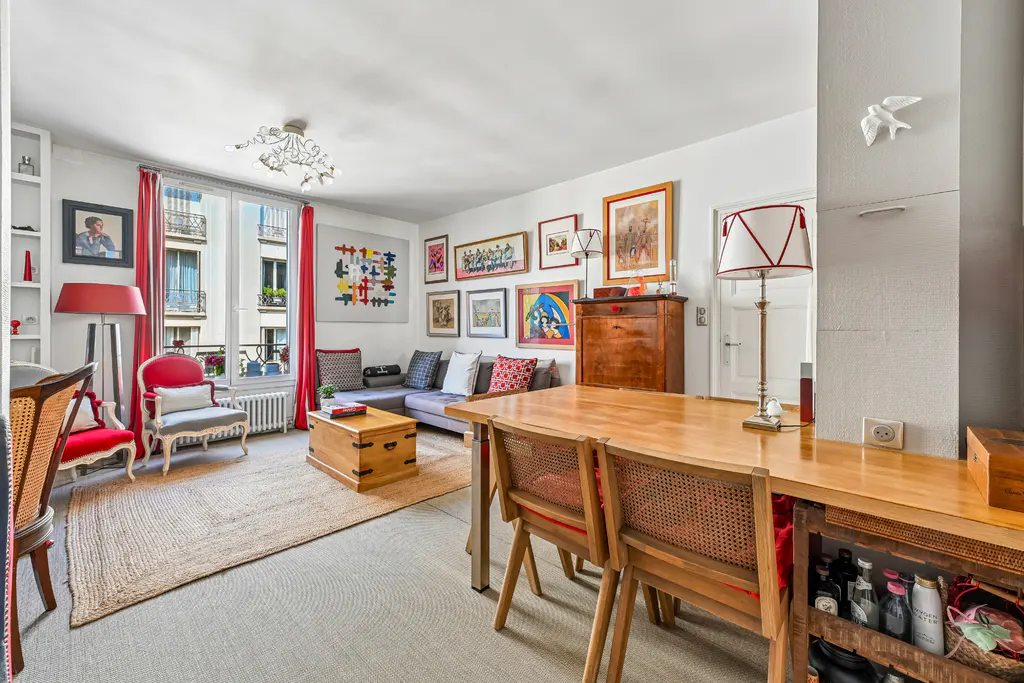 Bright living room with a gray sofa, wooden table, and red accents. Art adorns the walls, and a window offers a view of a building exterior.