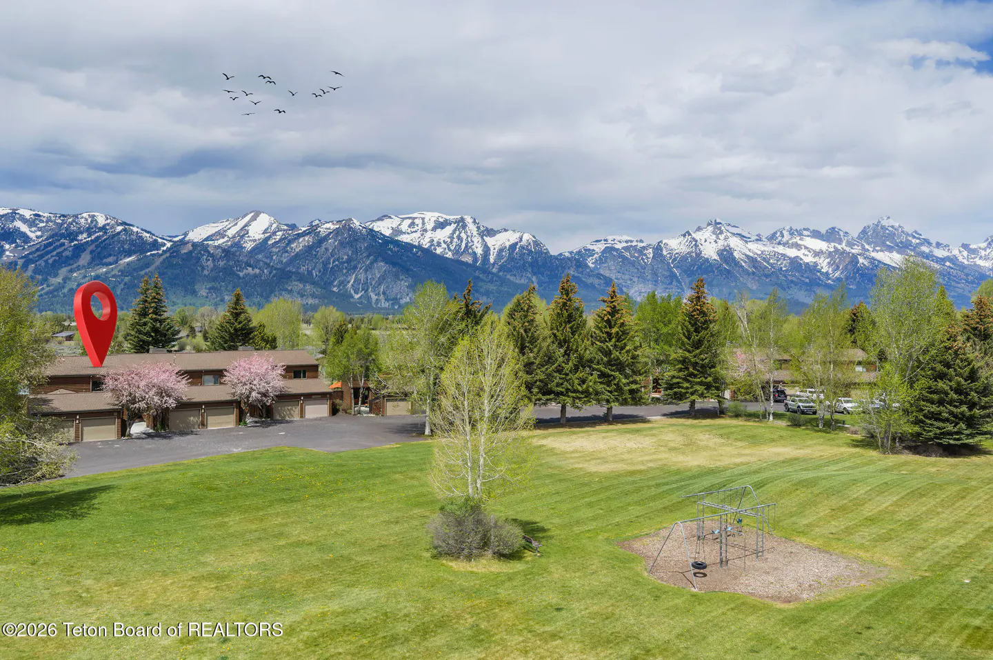 Exterior view of a brown townhouse with a red location pin, green lawn, playground, and snow-capped mountains in the background.