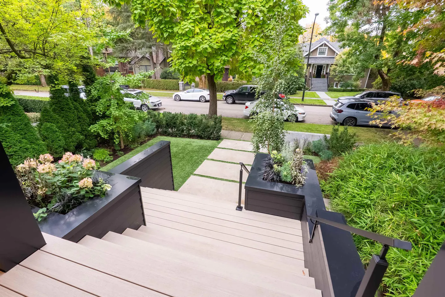 View from the top of light wood stairs with black railings, leading to a green lawn and street with parked cars.