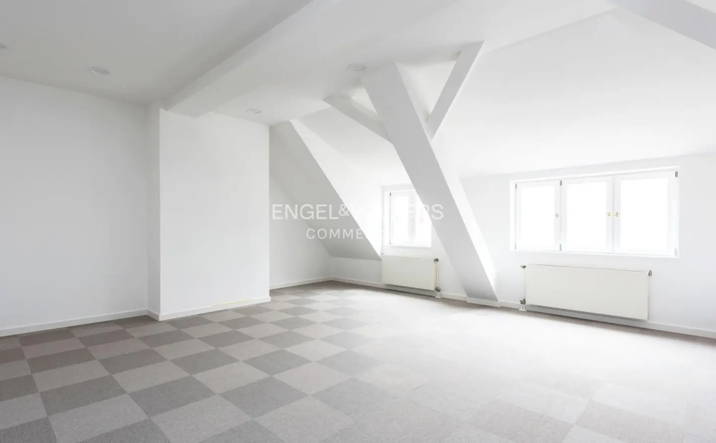 Bright, empty room with white walls, beams, and radiators. Gray and white checkered carpet. Two windows let in natural light.