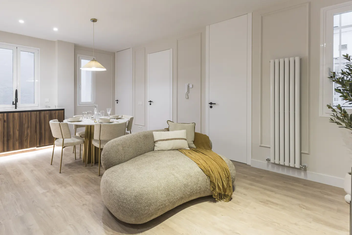 Modern open-plan living area with a beige curved sofa, dining table, and kitchen with wood cabinets and white walls.