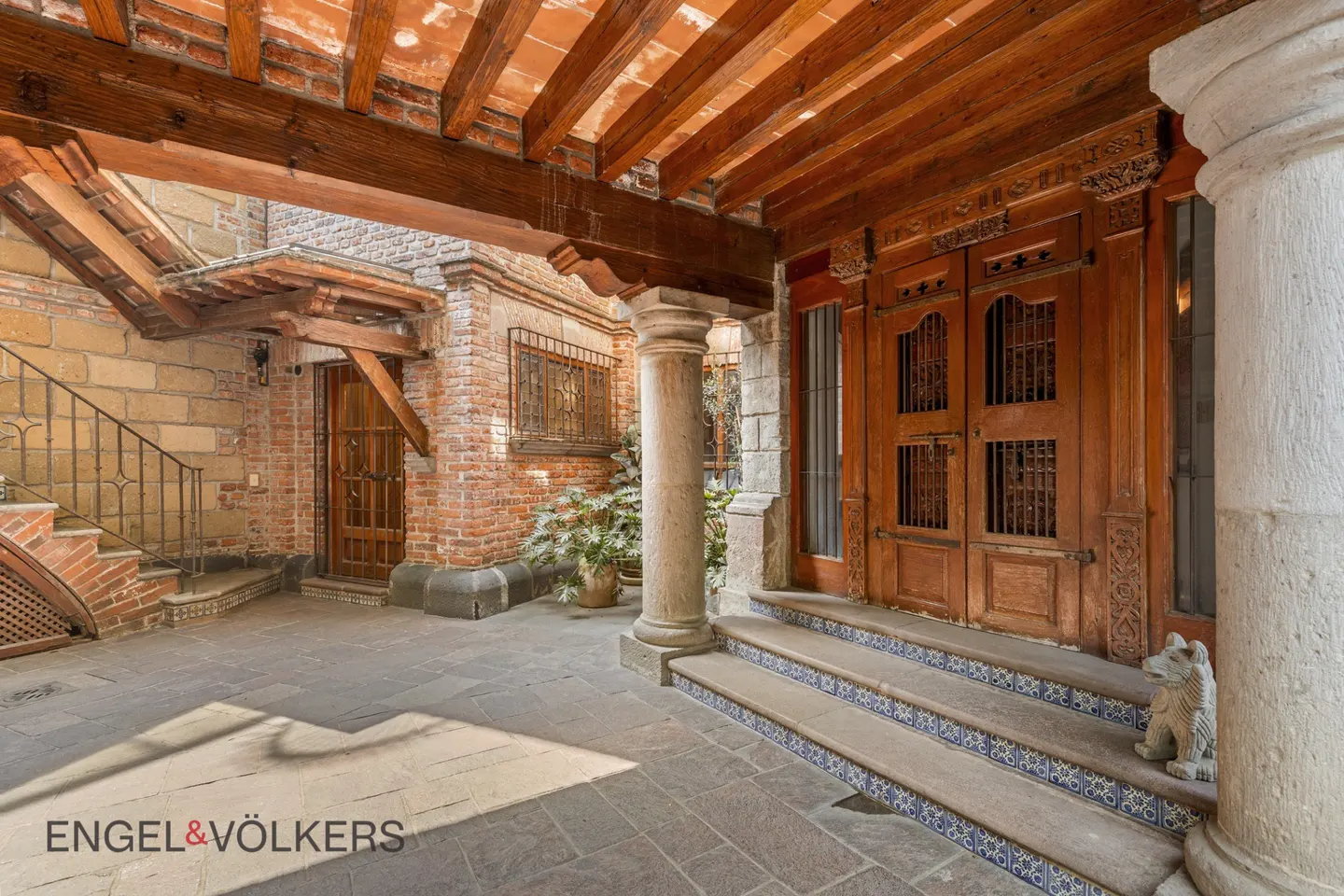 Exterior view of a courtyard with stone floors, brick walls, and a wooden ceiling with exposed beams. A large wooden door is visible on the right.