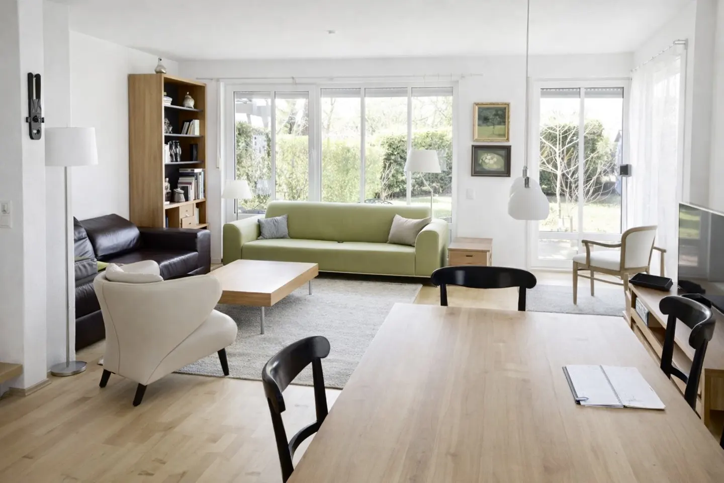 Bright living room with a green sofa, brown leather sofa, wooden table, and large windows overlooking a green garden.