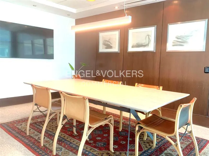 Conference room with a long white table, tan leather chairs, and a red patterned rug. Artwork hangs on the brown paneled wall.