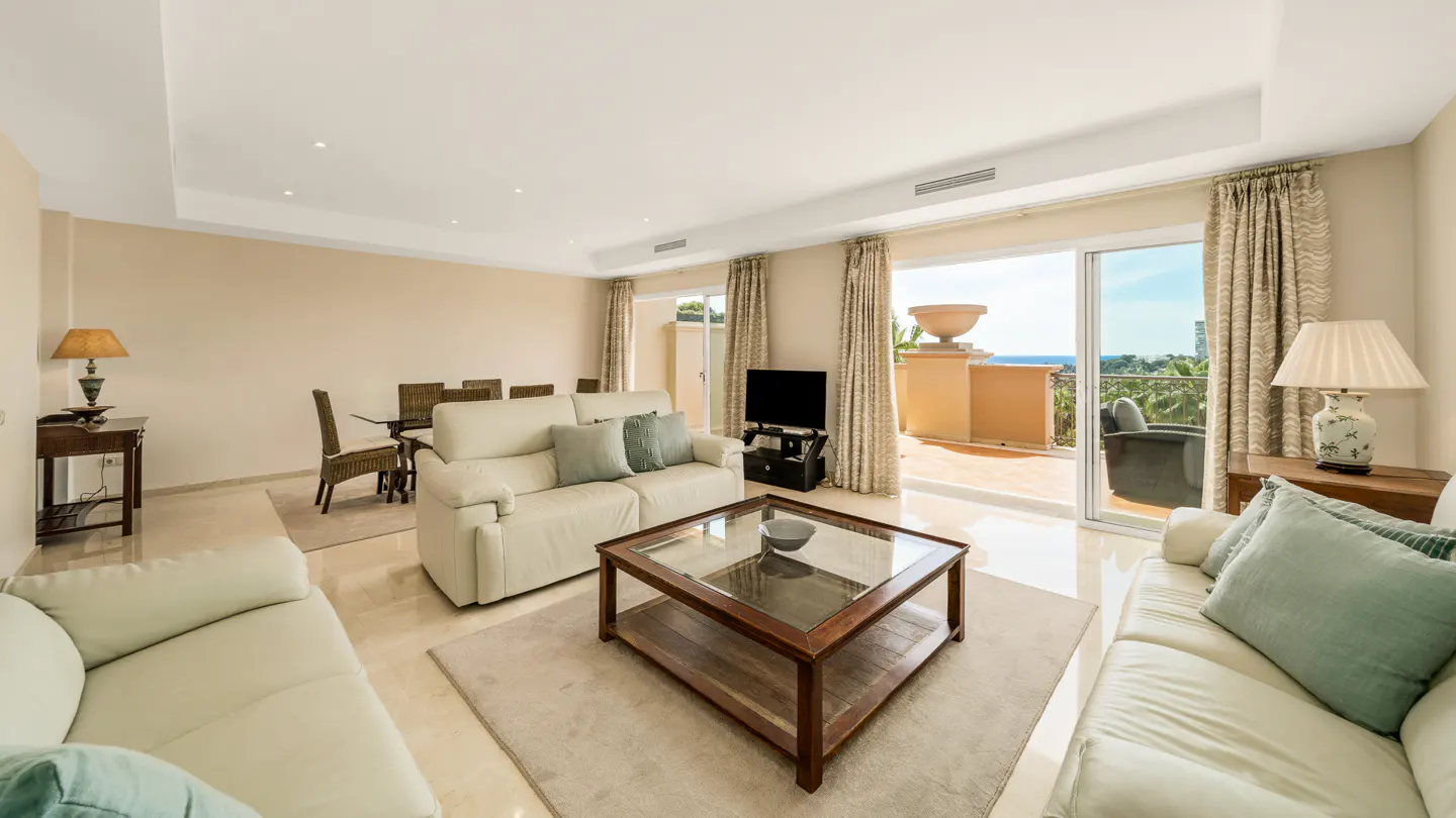Bright living room with beige walls, marble floors, and two white sofas. A glass coffee table sits on a light rug. Balcony with ocean view visible through sliding glass doors.