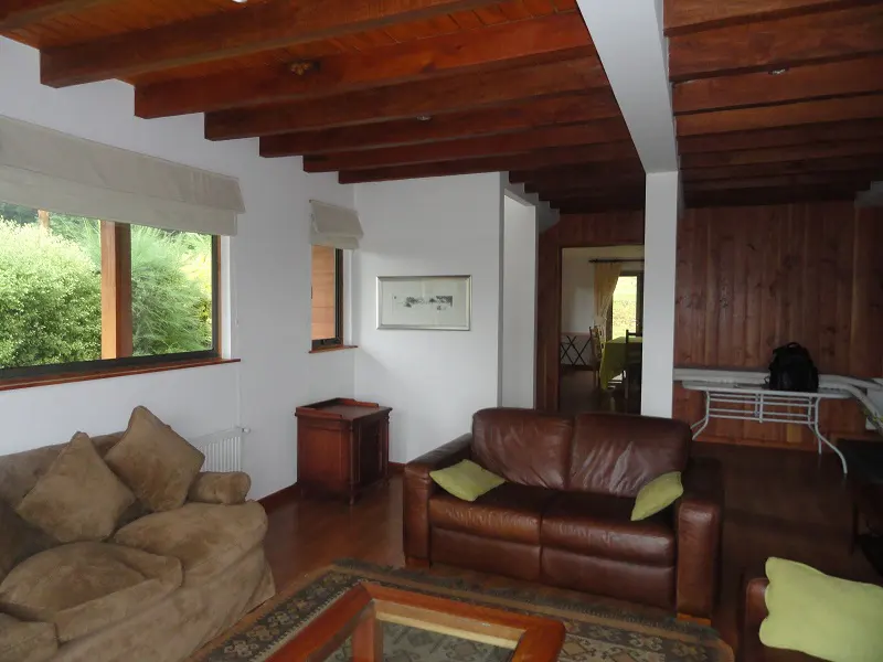 Living room with brown leather sofa, beige sofa, wood floors, and exposed wood beam ceiling. Windows overlook green foliage.