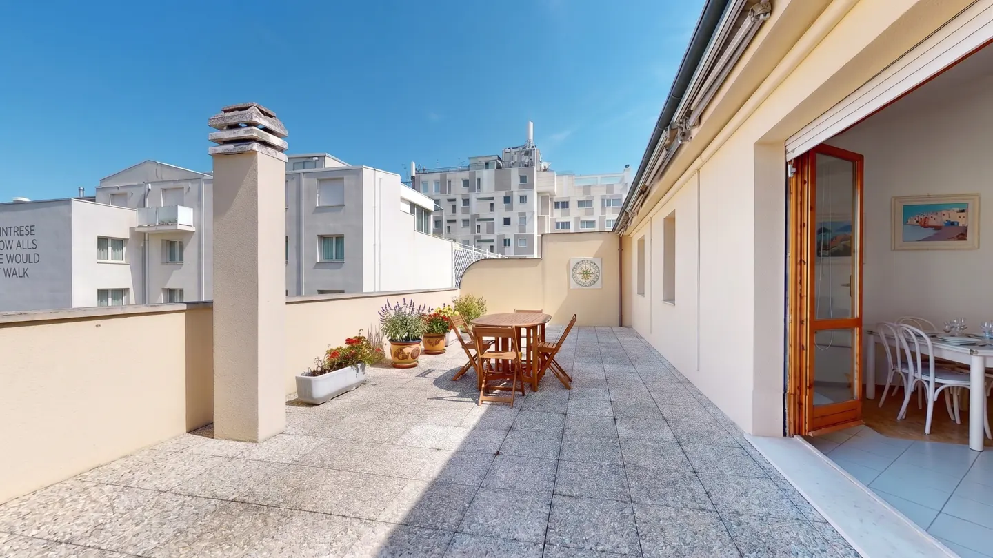 A sunny terrace with a table, chairs, and potted plants, leading to an open door with a dining room inside.