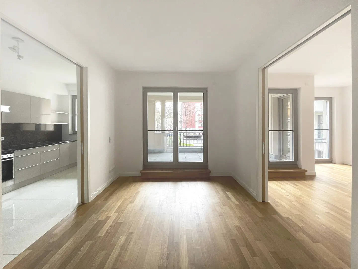 Bright, empty room with hardwood floors, white walls, and sliding glass doors to a balcony. A kitchen is visible through a doorway.