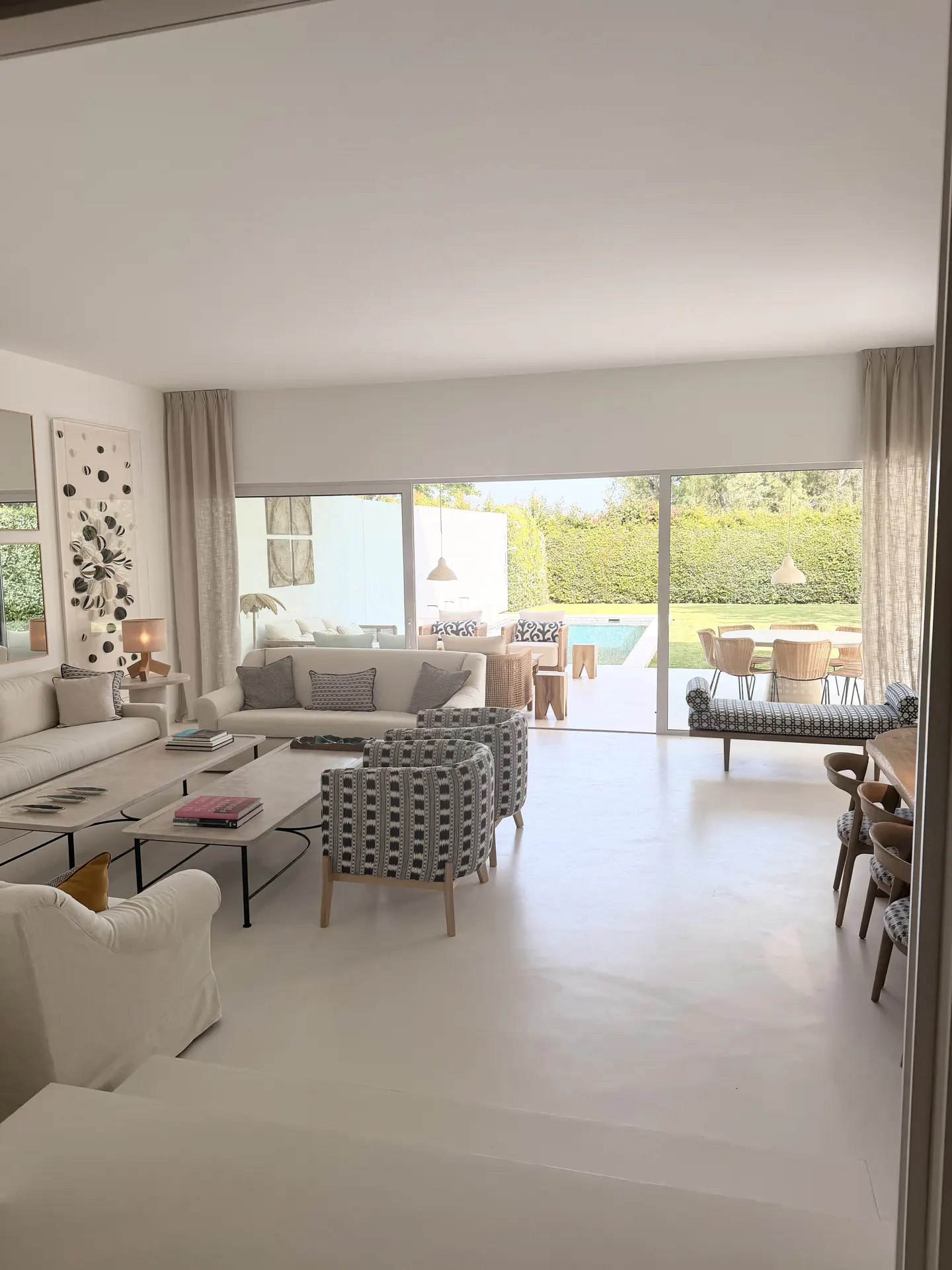 Bright, open living room with white sofas, patterned chairs, and large windows overlooking a pool and green lawn. Neutral tones create a serene atmosphere.