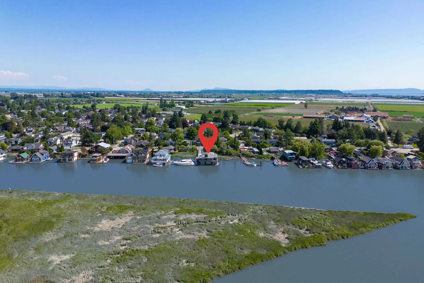 Aerial view of a house on the water, marked with a red pin, surrounded by greenery and a blue sky.