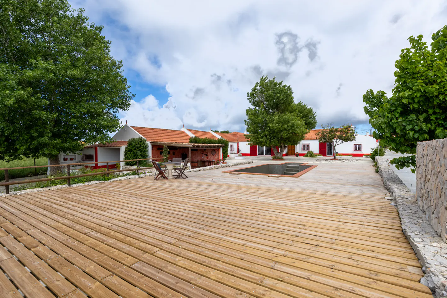 Exterior view of a property with a wooden deck, pool, and white buildings with red trim. Trees and cloudy sky in the background.