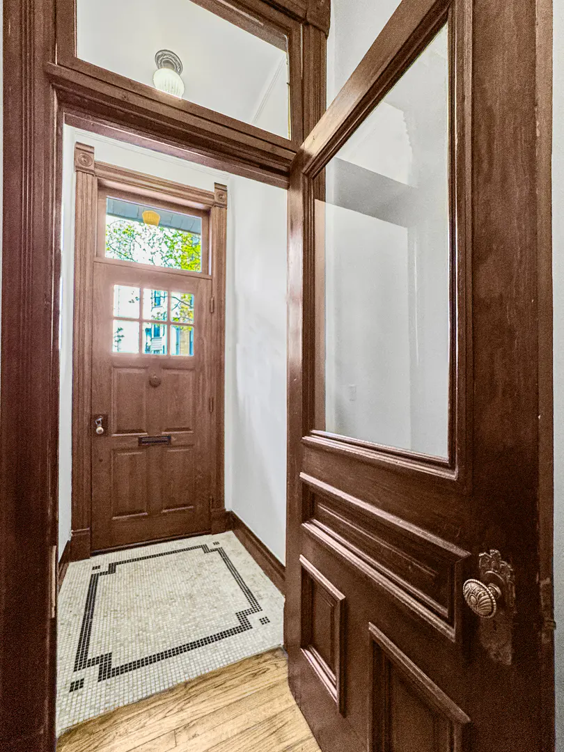 A brown front door with glass panes and a transom window, viewed from an open interior door. The floor has a mosaic tile rug.