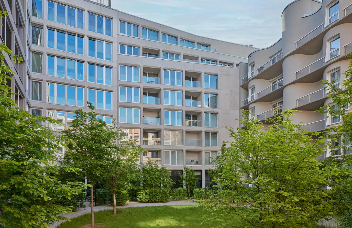 Modern apartment complex with large windows and balconies, surrounded by green trees and a grassy courtyard.