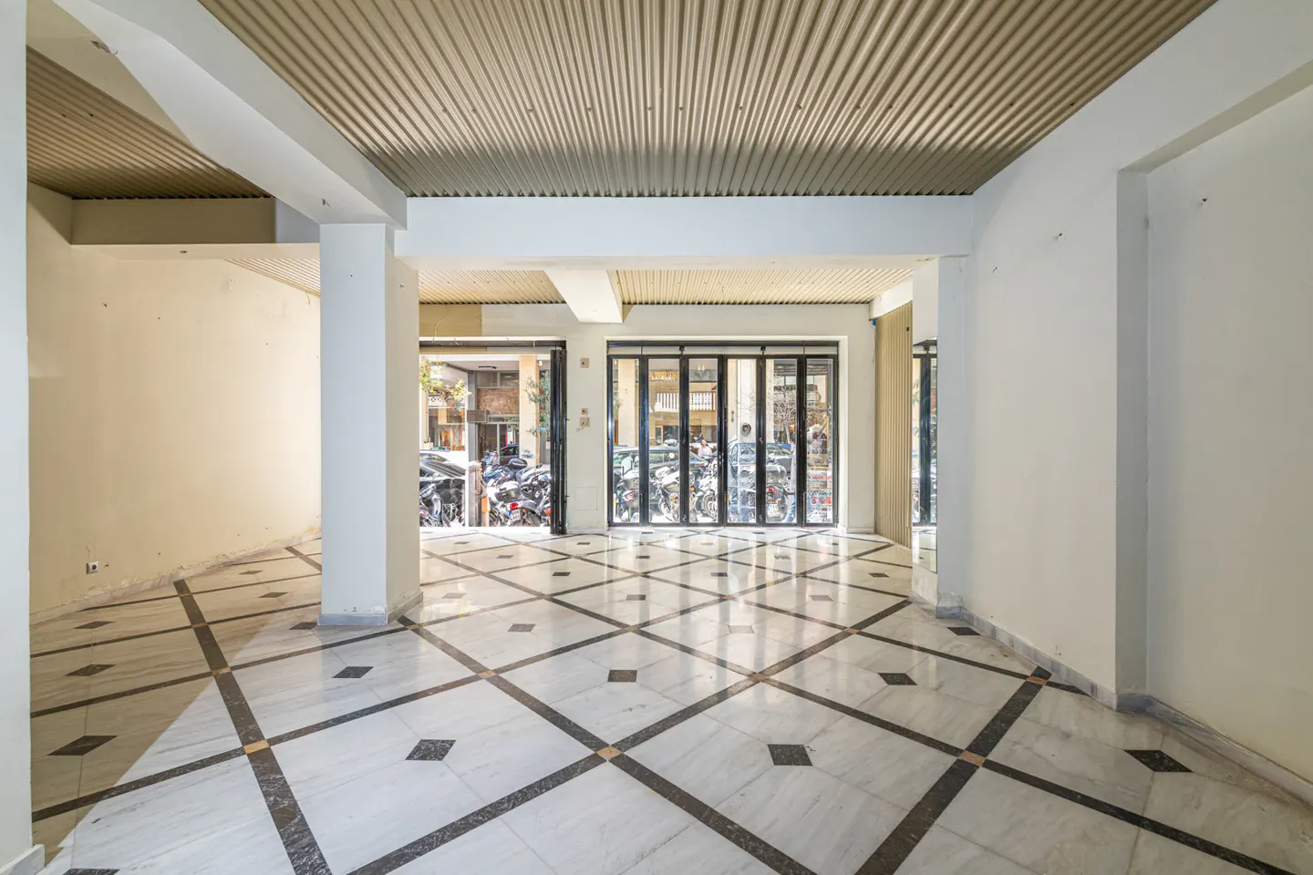 Empty commercial space with white walls, columns, and a patterned marble floor. Folding glass doors lead to a street with parked motorcycles.