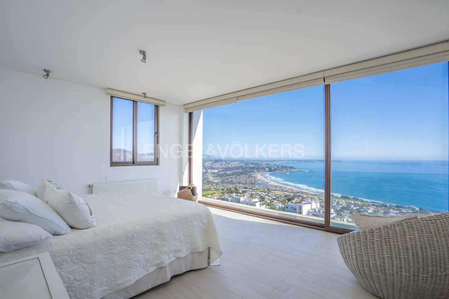 Bright bedroom with a white bed, large windows overlooking a coastal city and ocean view. Wicker chair in corner.