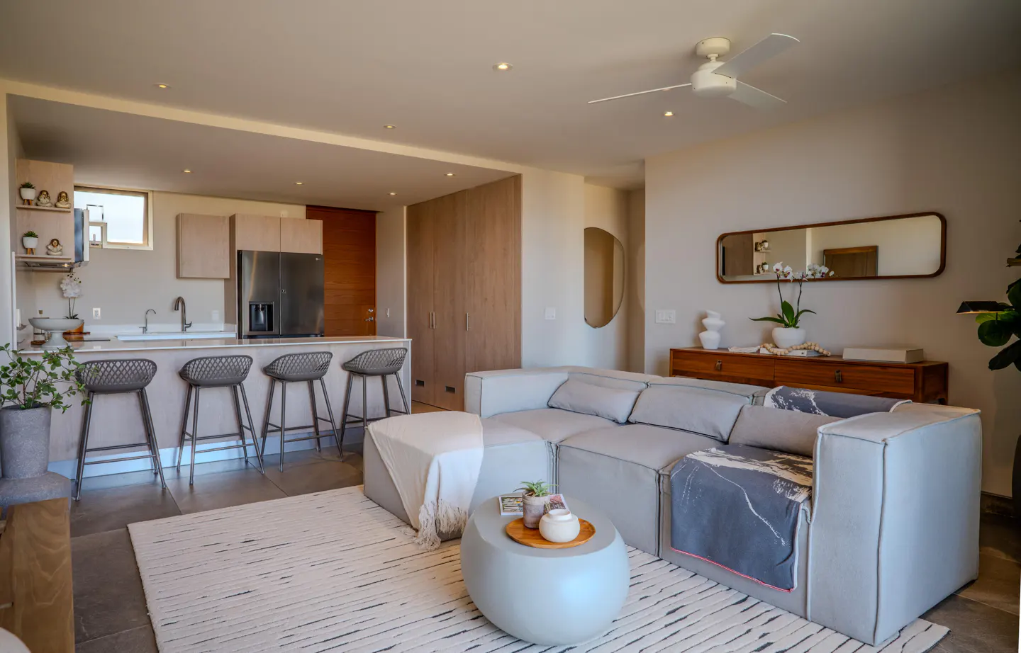 Open-concept living space with a gray sectional sofa, round coffee table, and kitchen island with four stools. Neutral color palette with wood accents.