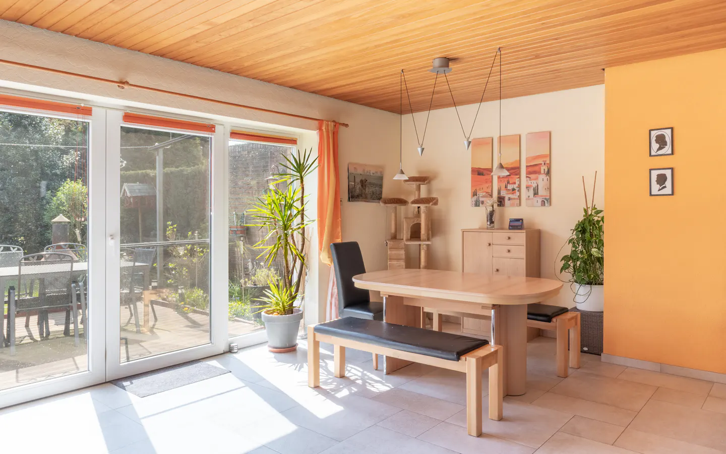 Bright dining room with wood ceiling, tile floor, table, bench, and chairs. Large glass doors lead to a patio and garden.
