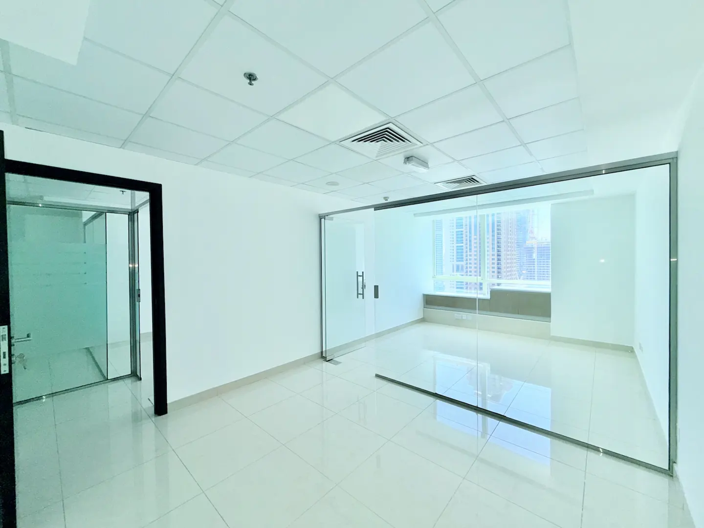 Bright, empty office space with white walls, tiled floor, and glass-walled rooms. A city skyline is visible through a window.