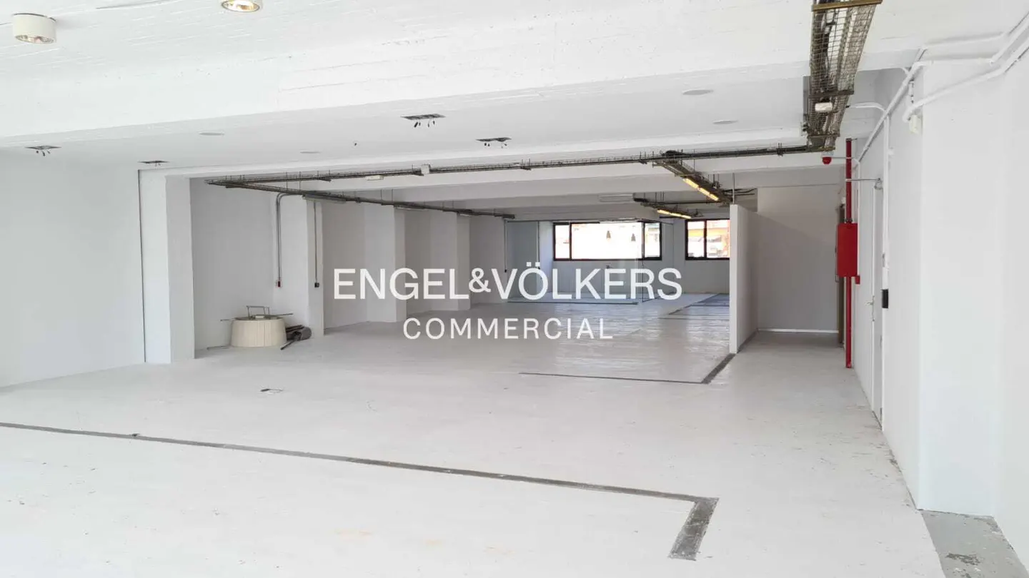 Bright, empty commercial space with white walls and floors. Overhead lighting and exposed pipes. Engel & Völkers Commercial logo overlay.