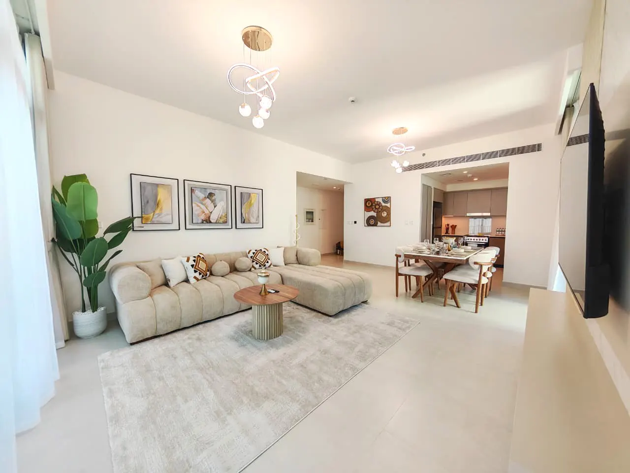 Bright, modern living room with beige sectional sofa, round wood table, and dining area with table set for six. Artwork adorns the walls.