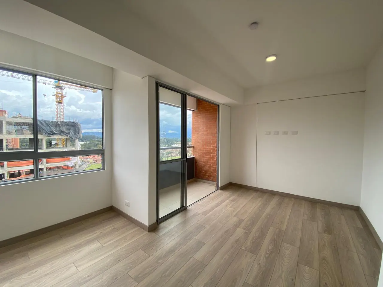Bright, empty room with wood floors, white walls, and a sliding glass door to a brick balcony. A large window shows a construction site.
