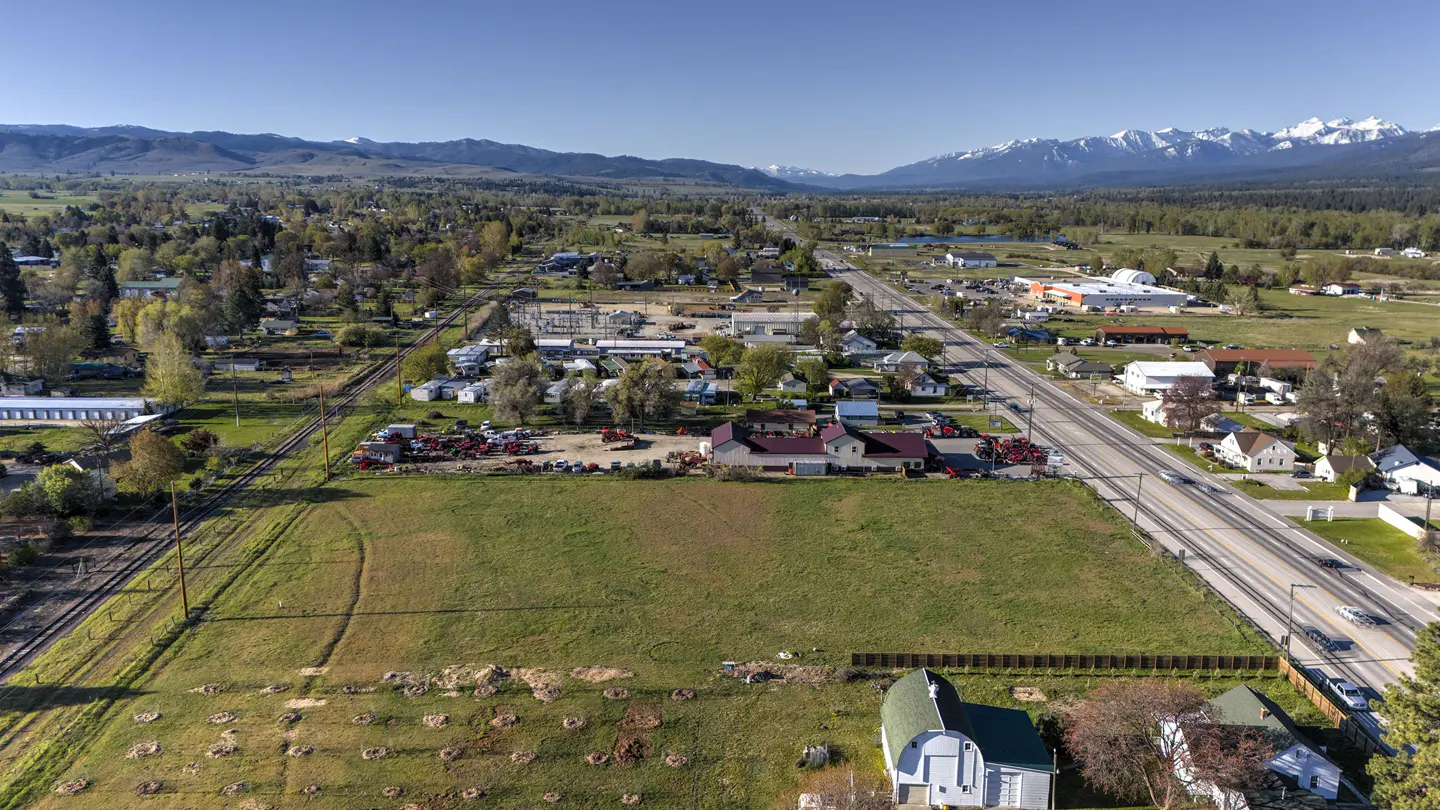 Aerial view of a rural town with a large grassy field, railroad tracks, and snow-capped mountains in the background.