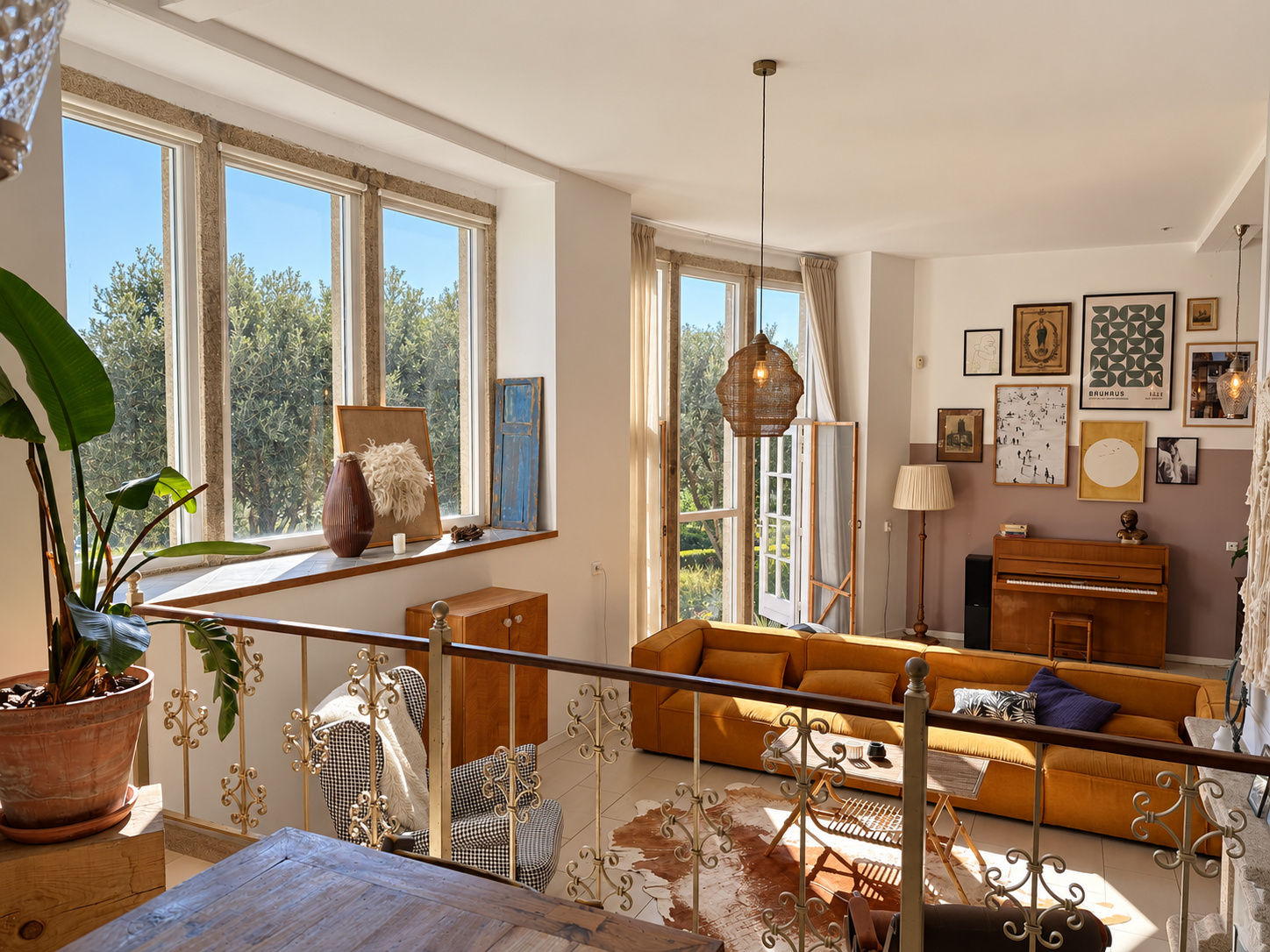 Sunlit living room with a mustard sofa, piano, gallery wall, and large windows overlooking trees, viewed from a balcony. [AI-generated text]