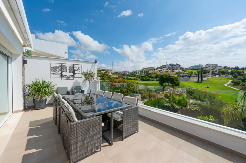 Outdoor patio with a glass table, wicker chairs, and a view of a golf course and blue sky.
