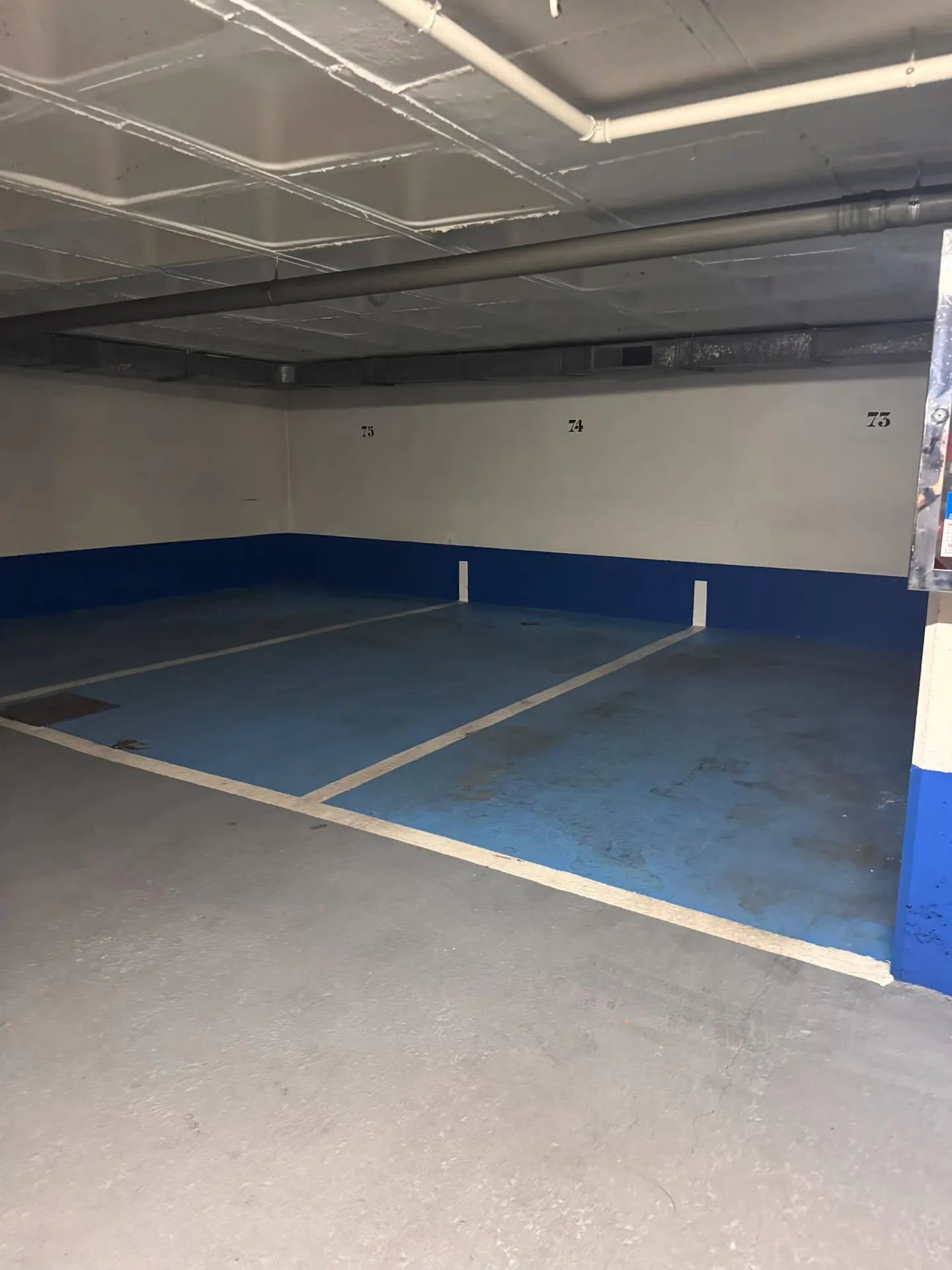 Empty indoor parking garage with blue painted floor spaces numbered 73, 74, and 75 against a white wall.