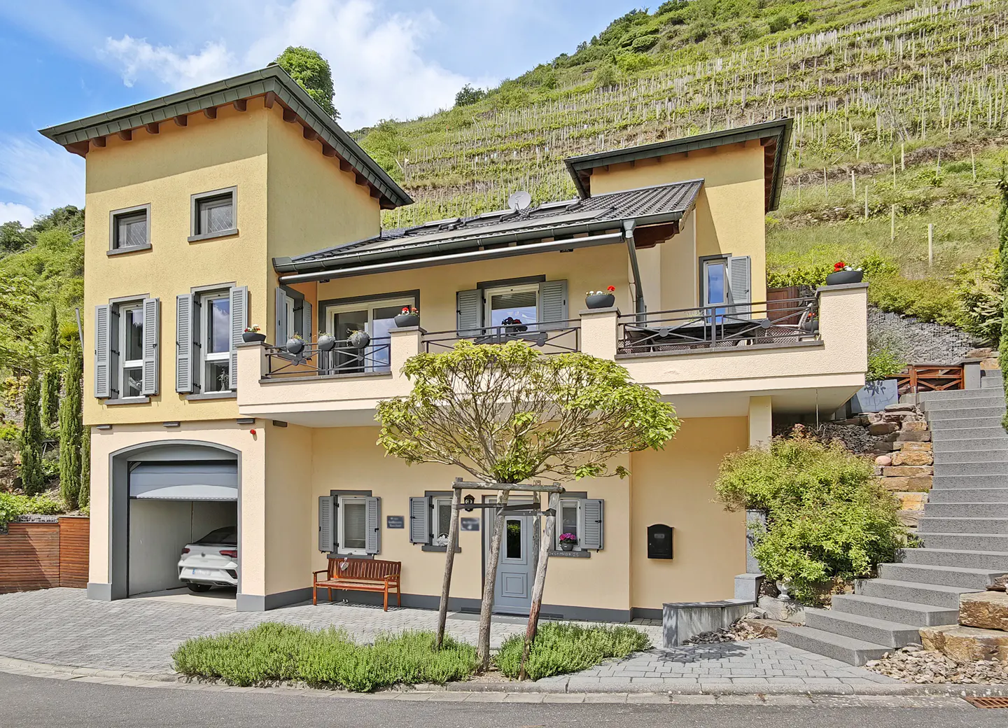 Yellow two-story house with a garage, balcony, and stone stairs, set against a terraced hillside vineyard.
