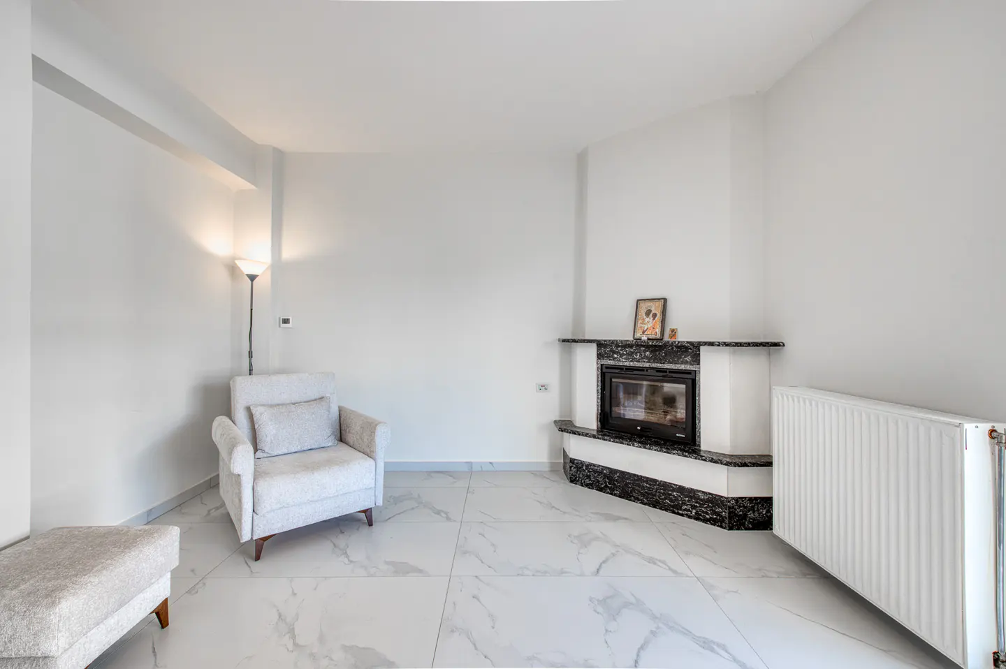 Bright living room with white walls, marble floors, and a black marble fireplace. A white armchair and ottoman sit near a floor lamp.