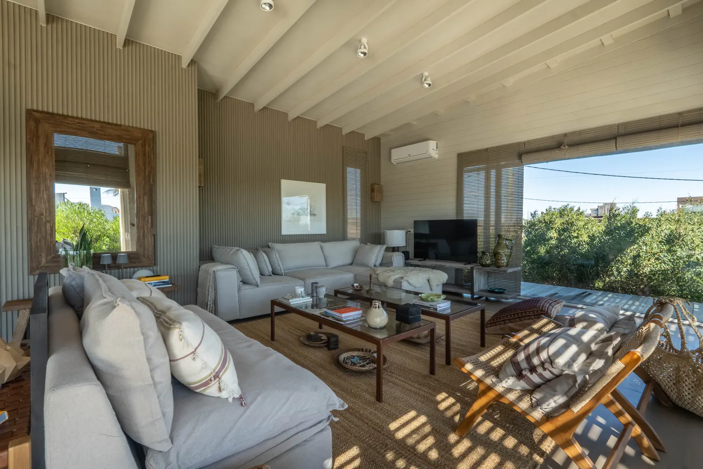 A bright living room with gray sofas, wooden tables, and a woven rug. A large window shows green trees outside.