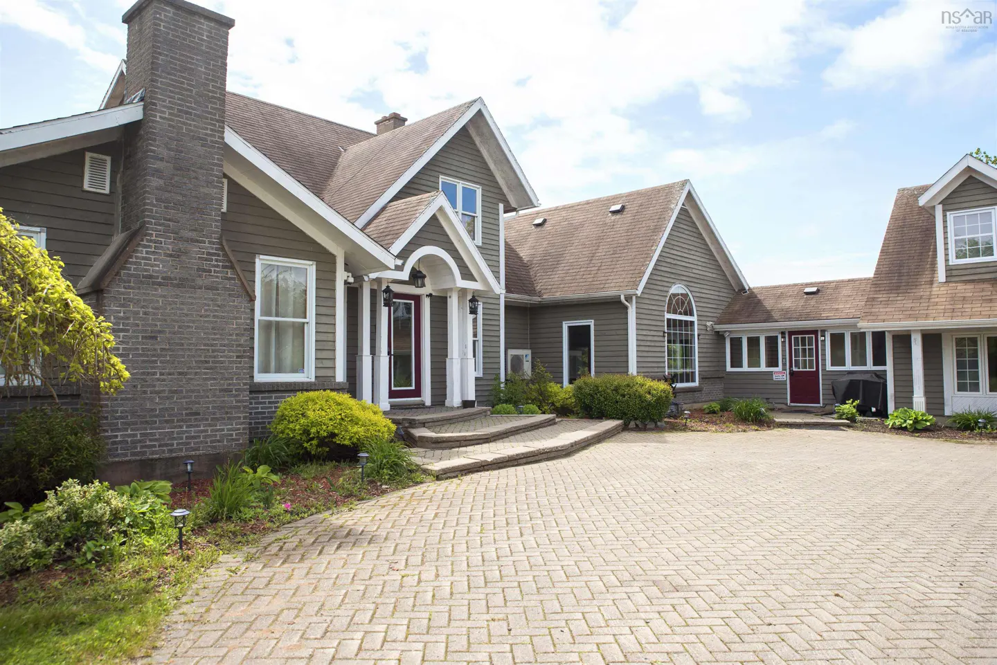 Exterior view of a large, two-story house with a brick driveway and a brick chimney. The house is painted in a neutral color with white trim.