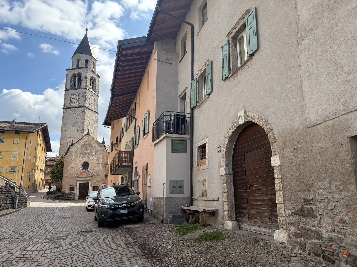 Cobblestone street view of a church tower and buildings in a European town. A dark car is parked on the side of the road.