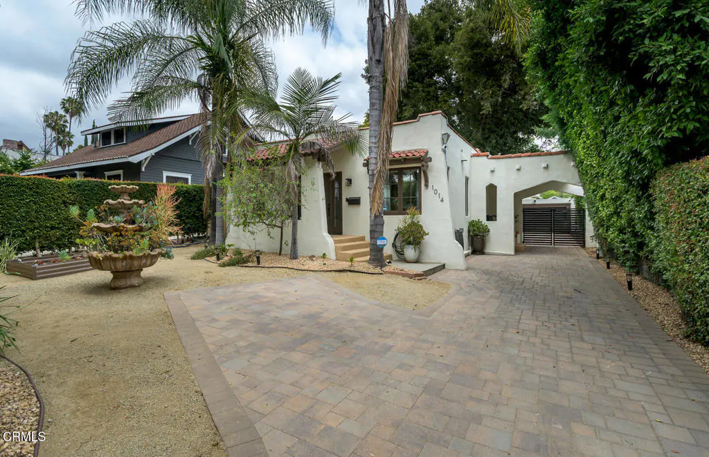 Exterior of a white stucco house with a red tile roof and a brick driveway. Palm trees and green hedges surround the property.