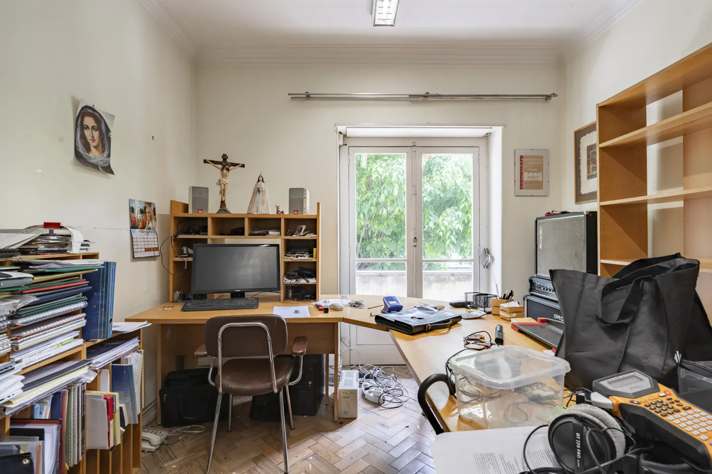 Cluttered office with a computer desk, chair, shelves, and a window view of green trees. Religious items are displayed.