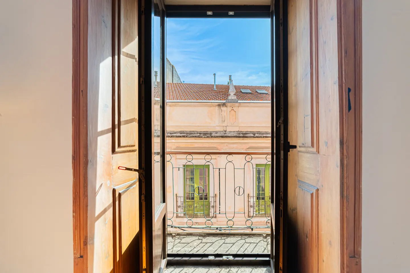 View through open wooden doors to a balcony with green doors and a building with a red tile roof under a blue sky.