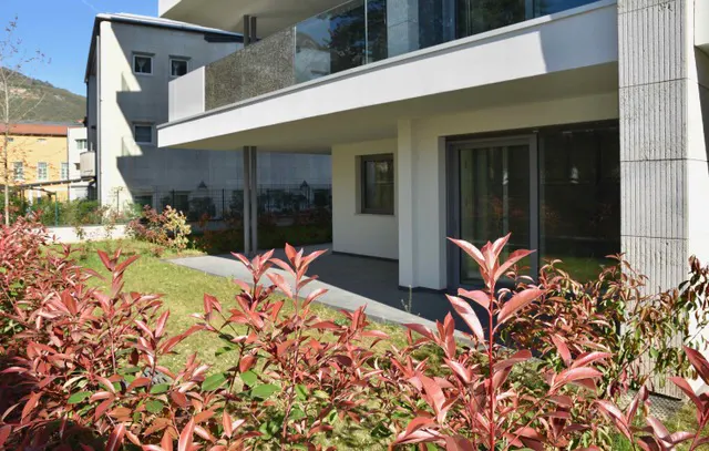 Modern white building with a covered patio and red-leaved bushes in the foreground.