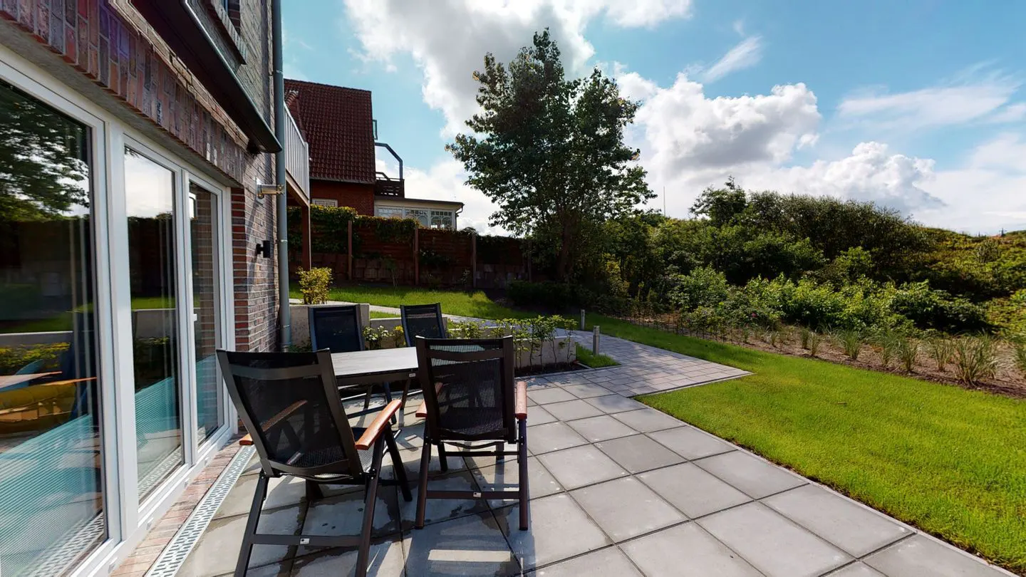 Outdoor patio with table and chairs on stone tiles, next to a brick building and green lawn. Blue sky with clouds above.