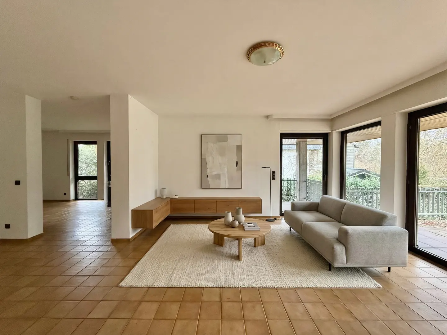 Bright living room with tan tile floor, white walls, and a gray sofa. A round wood table sits on a cream rug. A wood cabinet is against the wall.
