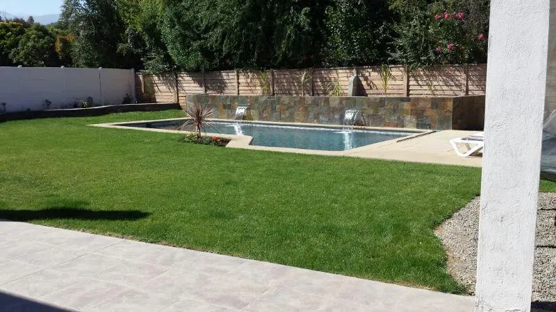 A backyard with a rectangular pool, green grass, and a stone waterfall feature. A white fence and trees are in the background.