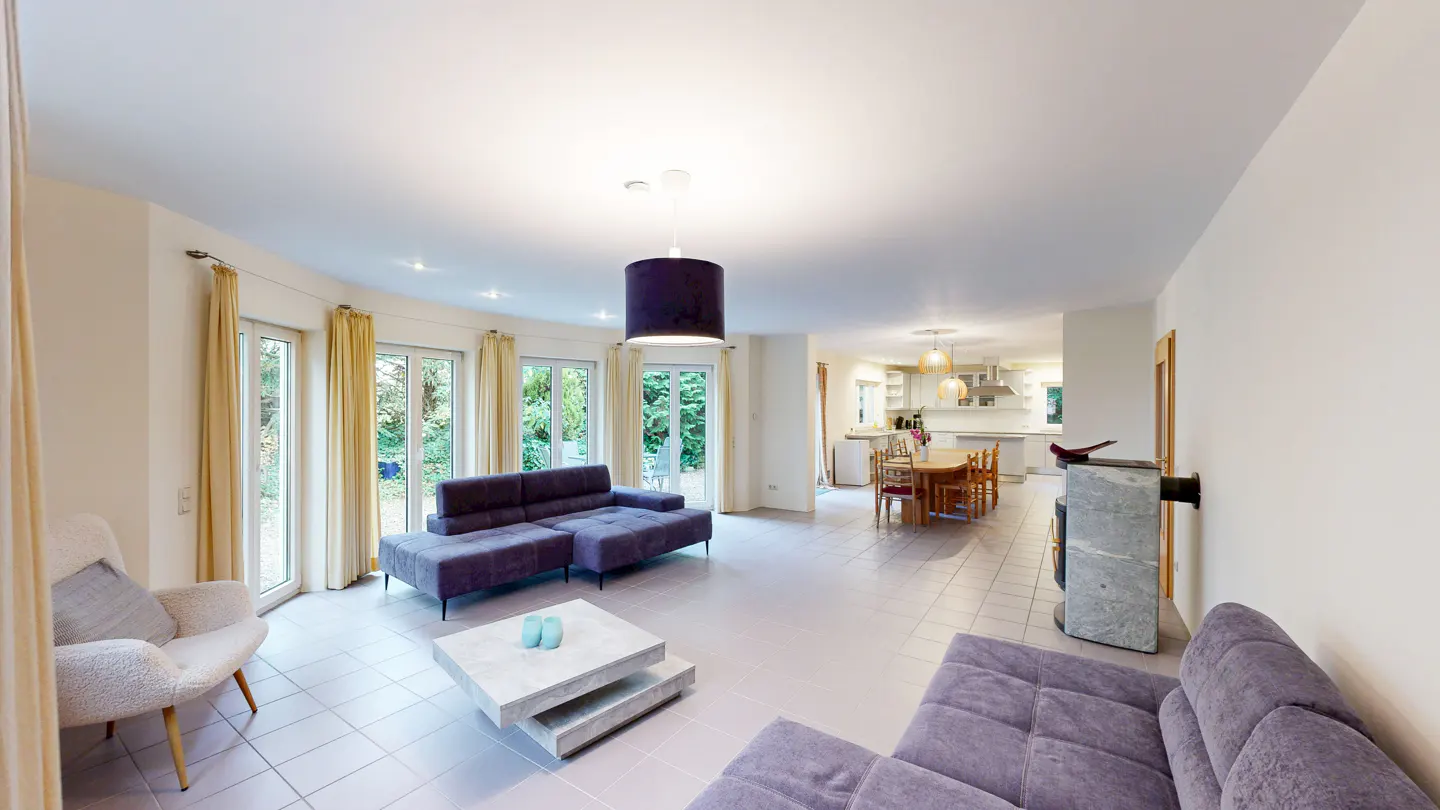Bright, open-concept living room with purple sofas, white walls, and tile floors. Large windows with yellow curtains overlook a green yard. Kitchen visible in the background.