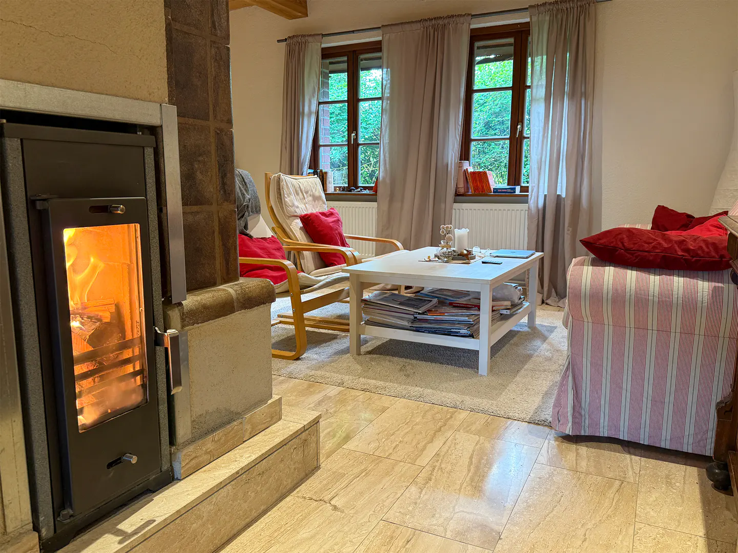 Cozy living room with a burning fireplace, two chairs with red pillows, and a white coffee table near the window.