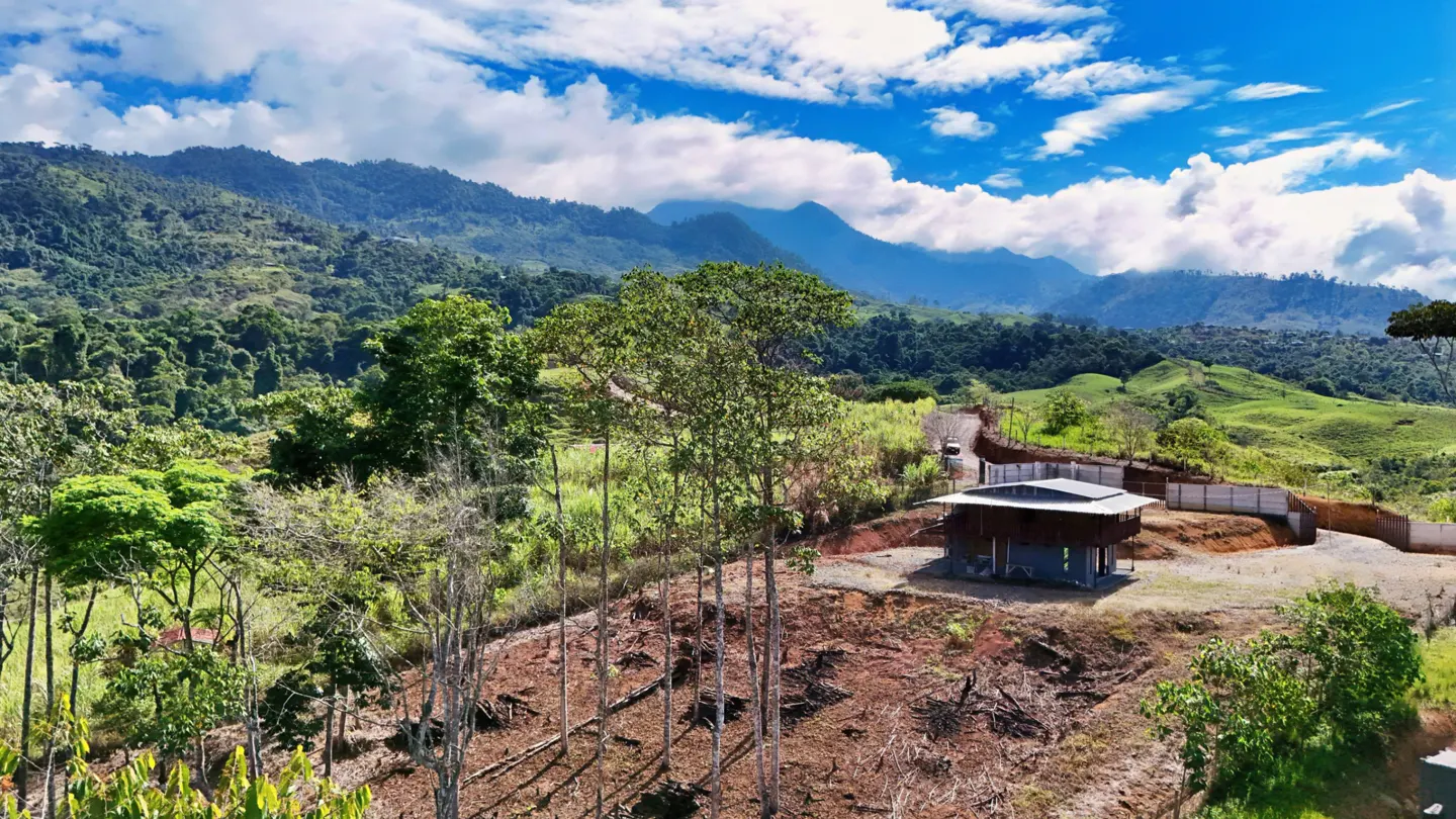 Scenic view of a modern house on a hillside, surrounded by lush green mountains under a blue sky with white clouds.