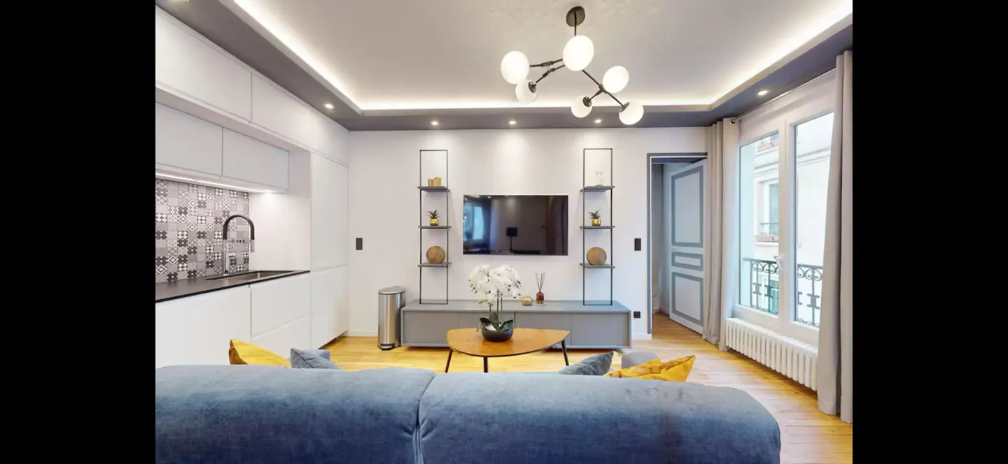 Bright living room with blue sofa, white walls, and modern lighting. A TV is mounted between two black shelves. A window lets in natural light.