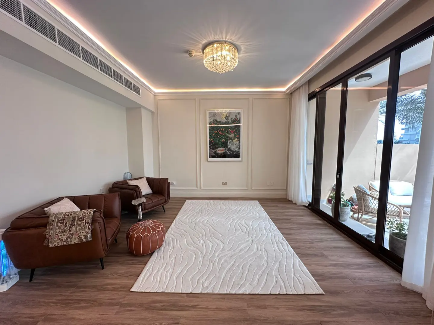 Living room with brown leather sofas, white rug, and sliding glass doors to a balcony. Chandelier and artwork add elegance.